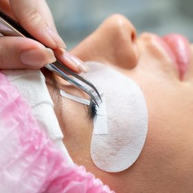 Eyelash extension master with tweezers in his hands performs eyelash correction close-up. Girl's eye with black artificial eyelashes closeup. Eyelash extension procedure in beauty salon. Lashmaker at work. Concept spa lash.